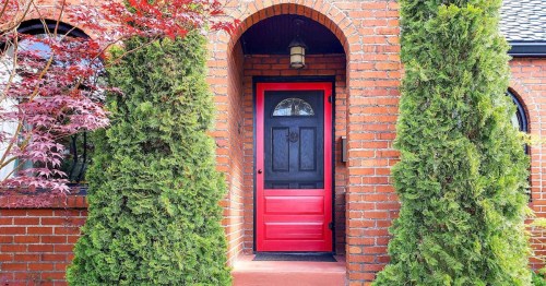 A red door framed by tall green shrubs and a brick archway, set against a brick wall.