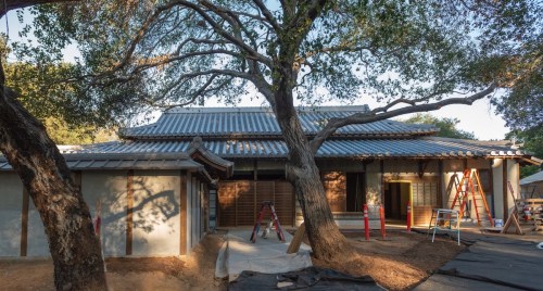 A traditional-style building under construction, surrounded by trees and scaffolding, with a tiled roof and open entrance.