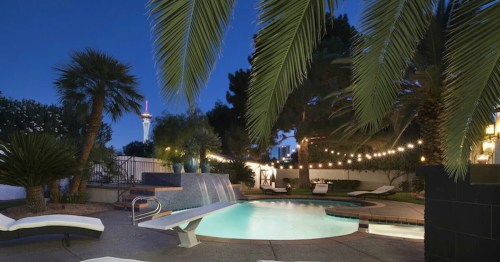 A serene pool area at night, surrounded by palm trees and string lights, with a view of a tower in the background.