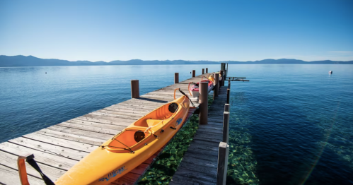 A wooden dock extends over clear blue water, with yellow kayaks lined up along the side and mountains in the background.