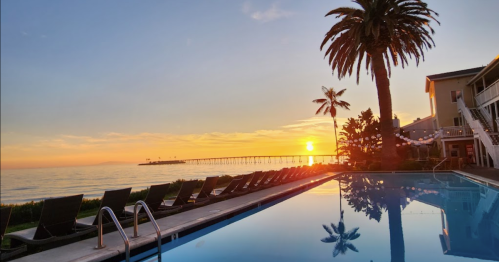 Sunset over a calm ocean, reflecting in a pool with palm trees and lounge chairs nearby.