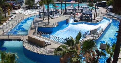 Aerial view of a water park featuring a lazy river, wave pool, and lounge areas with palm trees.