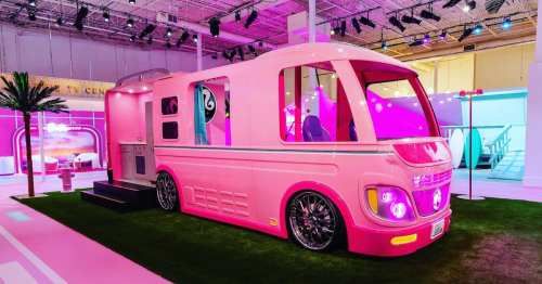 A vibrant pink retro-style camper van with large windows, set on green grass in a colorful exhibition space.