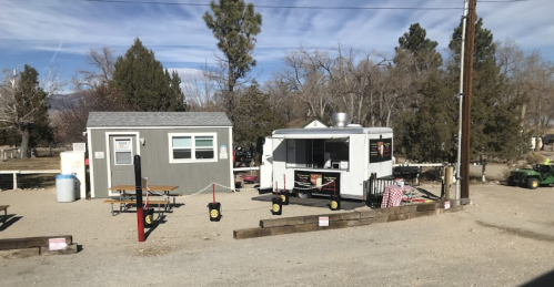 A food truck and a small building in a gravel area, surrounded by trees and a clear blue sky. Picnic tables are visible.