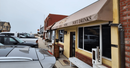 A storefront with a sign for soft drinks, wine, and beer, surrounded by parked cars on a cloudy day.