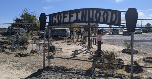 Sign reading "Coffinwood" at a fenced area with sparse vegetation and vehicles in the background.
