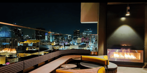 A rooftop lounge with modern seating and a fireplace, overlooking a vibrant city skyline at night.