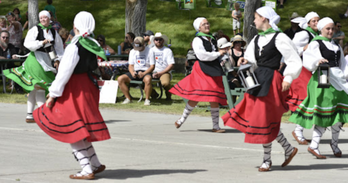A group of dancers in traditional costumes perform at an outdoor event, with spectators seated in the background.