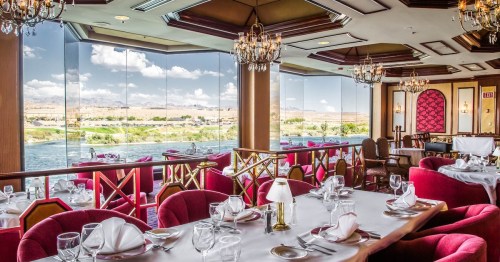 Elegant restaurant interior with plush red seating, chandeliers, and large windows showcasing a scenic view.