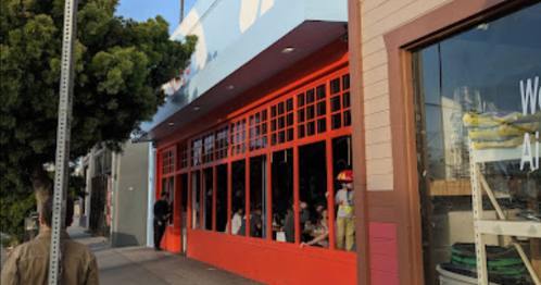 A colorful restaurant exterior with large windows, people dining inside, and a tree nearby on a sunny day.