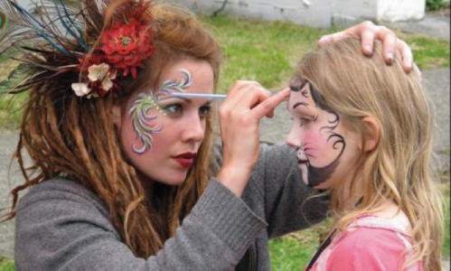 A woman applies face paint to a young girl, creating colorful designs on her face in an outdoor setting.