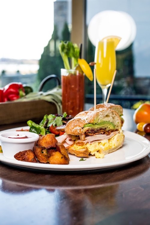 A delicious breakfast plate featuring a croissant sandwich, potatoes, salad, and colorful drinks on a wooden table.