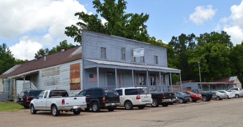 A weathered building with a sign, surrounded by parked cars, under a blue sky with scattered clouds.