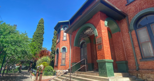 Historic red brick building with arched windows and a staircase, surrounded by trees and greenery under a clear blue sky.