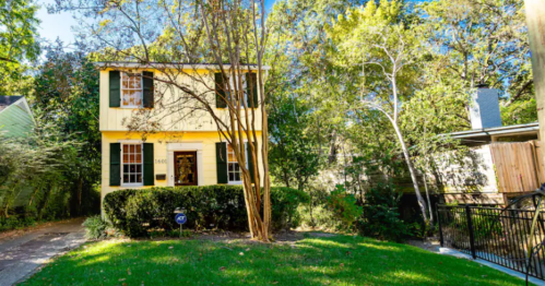 A two-story yellow house surrounded by greenery and trees, with a well-maintained lawn and a pathway.