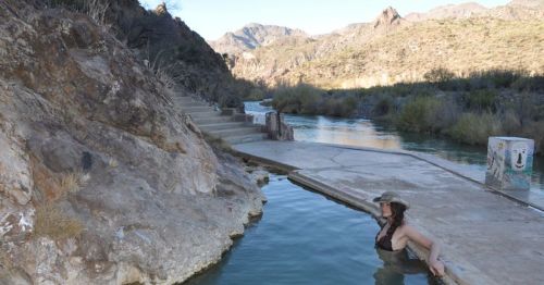 A woman relaxes in a natural hot spring surrounded by rocky terrain and mountains under a clear sky.