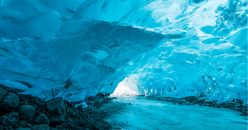 A stunning ice cave with blue hues, featuring smooth, glacial walls and a rocky floor leading to a bright exit.