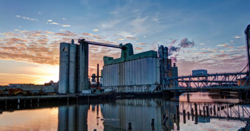 Industrial buildings by a river at sunset, with colorful clouds reflecting in the water.