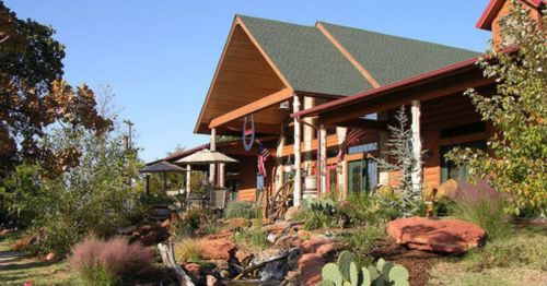 A rustic wooden house with a green roof, surrounded by landscaping, rocks, and cacti under a clear blue sky.