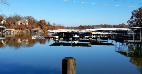 A calm lake scene with boat docks, reflecting trees and clear blue skies in the background.