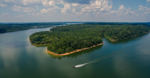 Aerial view of a lush green island surrounded by calm water under a partly cloudy sky. A boat is visible in the distance.