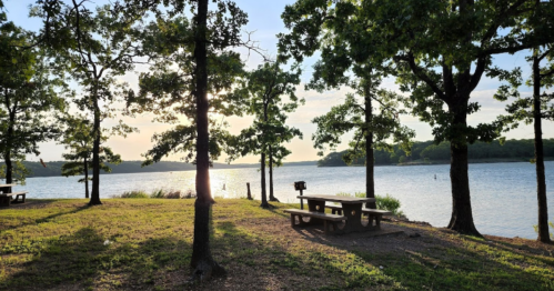 A serene lakeside scene with picnic tables, trees, and a sunset reflecting on the water.