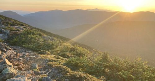 A scenic mountain landscape at sunset, with sun rays illuminating the peaks and lush greenery along the rocky path.