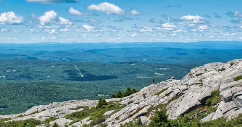 A panoramic view from a rocky mountain summit, showcasing lush green valleys and distant blue hills under a cloudy sky.