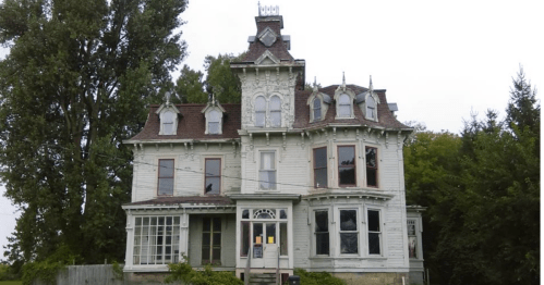 A large, Victorian-style house with intricate architecture, surrounded by trees and a cloudy sky.
