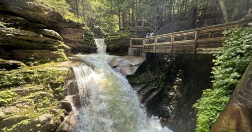 A scenic waterfall cascades through rocky terrain, surrounded by lush greenery and wooden walkways.
