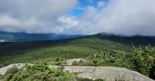 A panoramic view of lush green mountains under a partly cloudy sky, with rocky foreground and distant peaks.