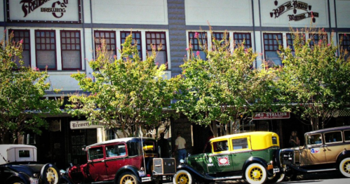 Vintage cars parked in front of a historic brewery building, surrounded by trees and a clear blue sky.
