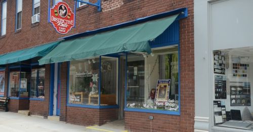 Exterior of a yarn shop with a green awning and a sign reading "Distinctive Knits Yarn Shop" on a brick building.
