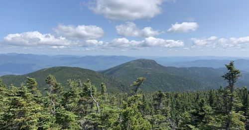 A panoramic view of green mountains under a blue sky with fluffy clouds, showcasing a serene natural landscape.