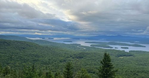 A panoramic view of lush green hills and a serene lake dotted with islands under a cloudy sky.