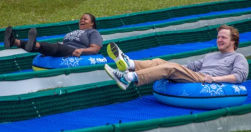 Two people joyfully sliding down a water slide on inflatable tubes, with green grass in the background.