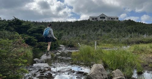 A hiker walks along a rocky path near a pond, with a house visible on a hill in the background under a cloudy sky.