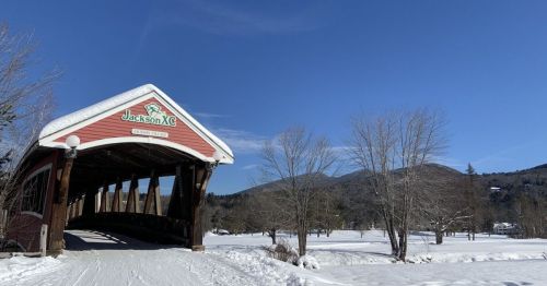 A snow-covered landscape featuring a red covered bridge with "Jackson XC" sign, surrounded by trees and mountains.