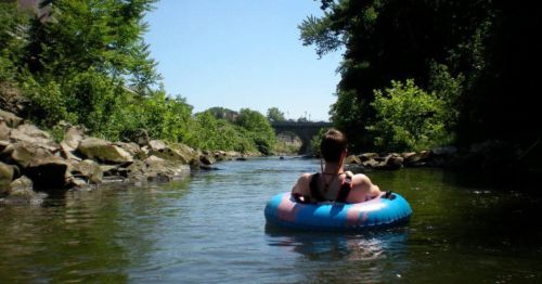 A person sits in a blue inner tube on a calm river, surrounded by greenery and a bridge in the background.