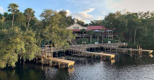 A serene view of wooden docks by a calm river, surrounded by trees and a building in the background under a blue sky.