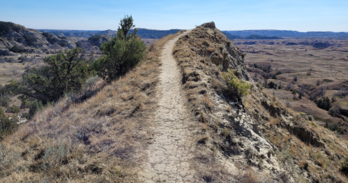 A narrow dirt path winds along a ridge, surrounded by rugged terrain and distant hills under a clear blue sky.