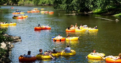 People floating on colorful inner tubes in a river, surrounded by lush greenery and a sunny sky.