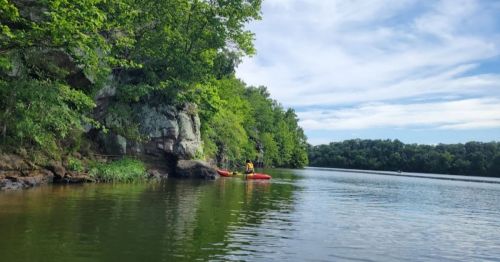 Two people in kayaks on a calm lake surrounded by lush greenery and rocky cliffs under a blue sky.