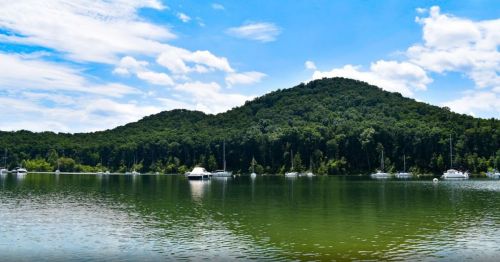 A serene lake surrounded by lush green hills and sailboats under a bright blue sky with scattered clouds.