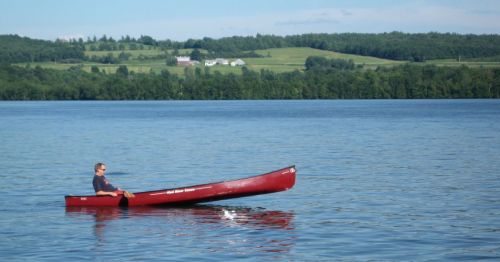 A person paddles a red canoe on a calm lake, surrounded by green hills and distant white houses.