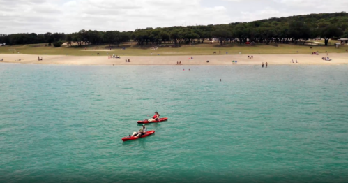 Two kayakers paddle on a turquoise lake, with a sandy beach and trees in the background.