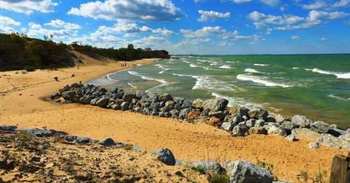 A sandy beach with rocky formations, gentle waves, and a blue sky dotted with clouds.