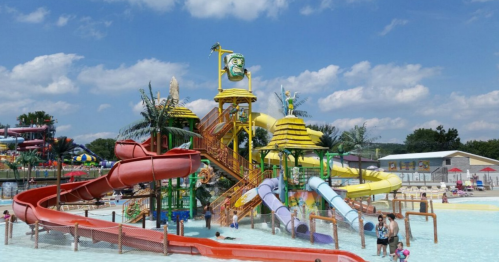 Colorful water park with multiple slides, pools, and palm trees under a blue sky with fluffy clouds. Families enjoying the water.