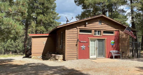 A rustic wooden cabin surrounded by tall pine trees, featuring a welcoming entrance and an American flag.