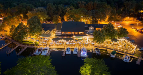 Aerial view of a lakeside restaurant with boats docked, surrounded by trees and illuminated by warm lights at night.